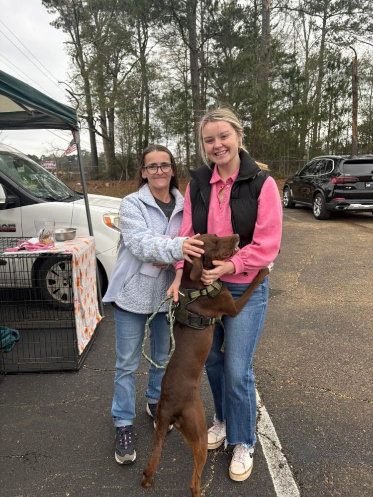 Two adopters smiling with their newly adopted brown rescue dog outside PALS Rescue in McComb, Mississippi